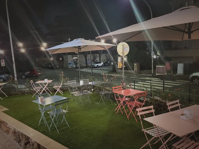 Dehors serale su prato sintetico con tavolini pieghevoli rosa, corallo e grigi, ombrelloni bianchi e luci appese sulla strada