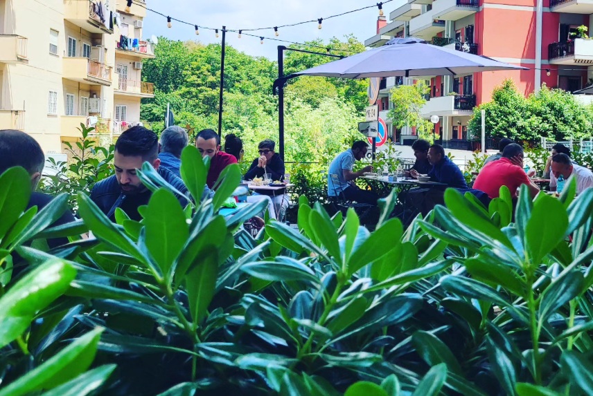 Terrazza di Casita Bar con clienti ai tavoli sotto un ombrellone grigio, lucine sospese, siepe verde e palazzi colorati